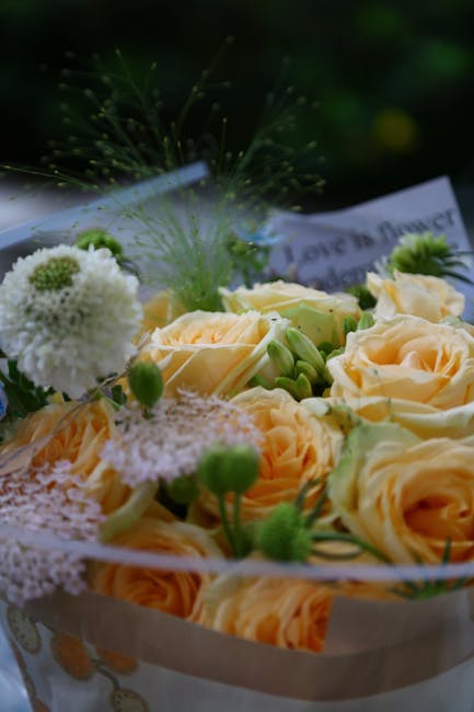 A close-up of a floral arrangement featuring soft peach-colored roses with fully open petals, surrounded by white pom-pom chrysanthemums and delicate green buds. The flowers are arranged in a white wr