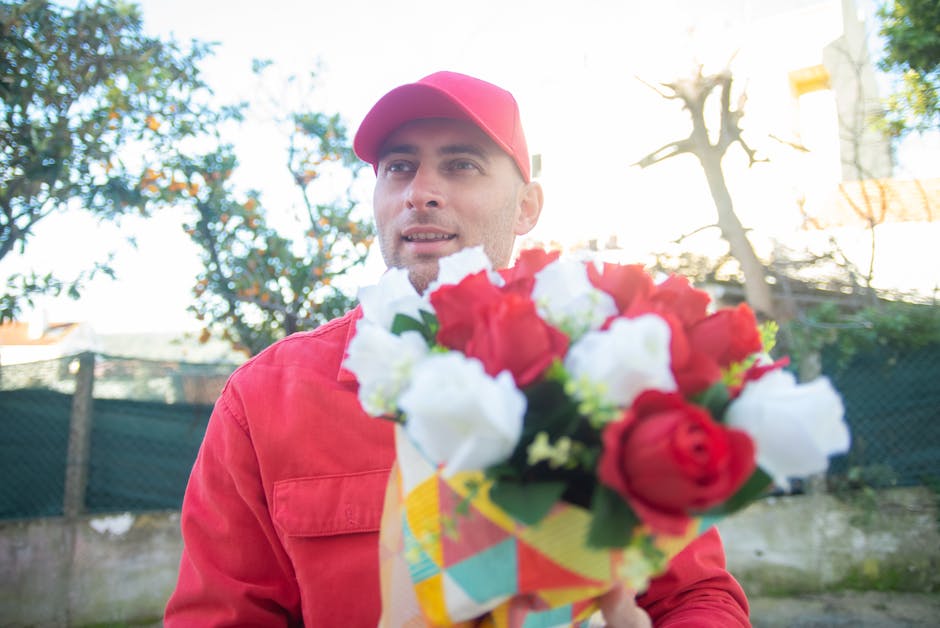 A man dressed in a red cap and red jacket holding a colorful bouquet of fresh flowers outdoors. The bouquet contains red and white roses along with green foliage, arranged in a decorative, multicolore