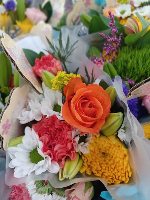 A vibrant floral arrangement featuring an orange rose as the central focal point, surrounded by white daisies, pink carnations, yellow chrysanthemums, and purple lavender sprigs. The flowers are wrapp
