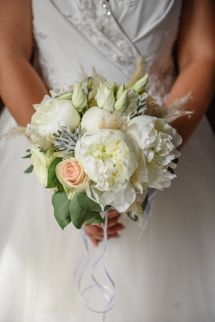 A bride holding a bouquet arrangement of fresh wedding flowers featuring predominantly white blooms such as peonies, roses, and lisianthus, accented with soft peach roses and silvery foliage. The bouq