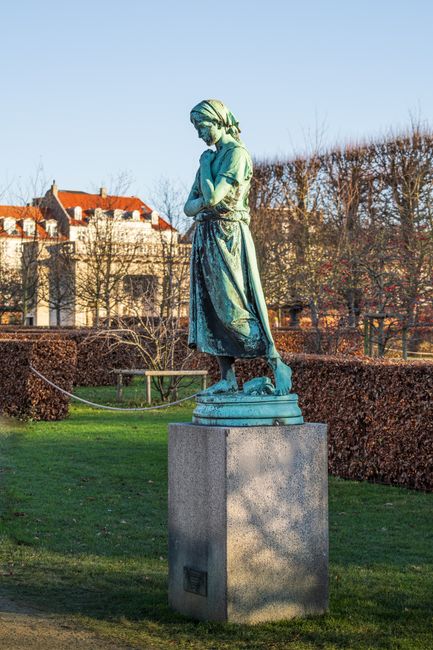 A bronze statue of a woman dressed in historical clothing, standing on a stone pedestal in a park setting. The statue depicts a female figure wearing a long skirt, apron, and head covering, with arms 